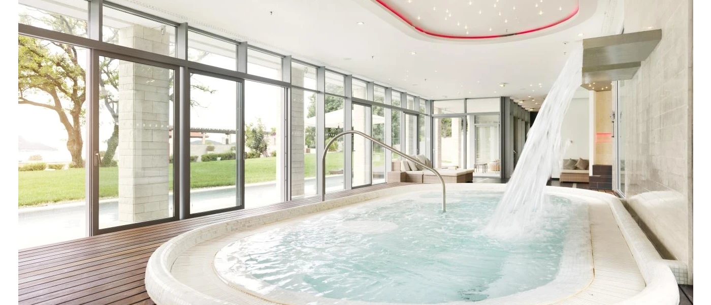 Indoor pool with water jets in wooden decking, with full-length windows showing the garden beyond