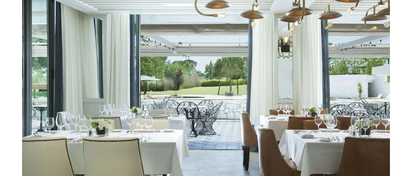 Stylish modern restaurant with white table cloths and curtains, black-framed patio doors, brass pendant lights and an outdoor terrace with black furniture