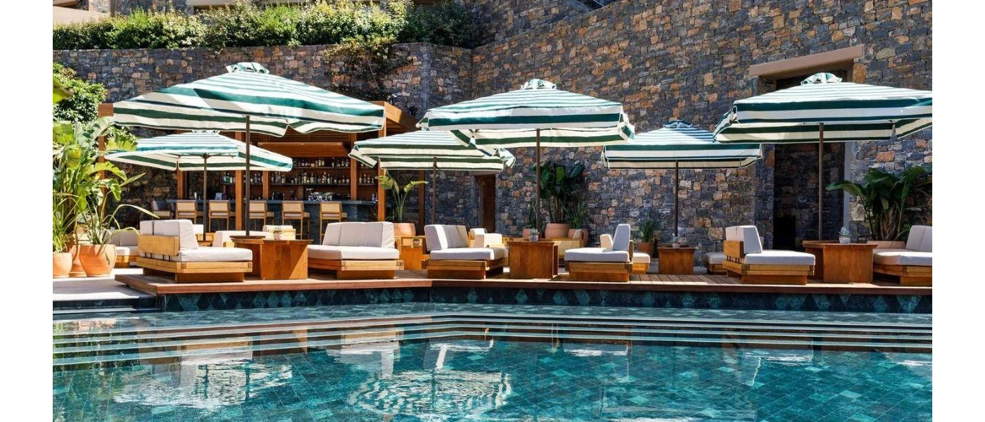Swimming pool with steps next to a sunny terrace with wooden white-cushioned loungers and green and white striped umbrellas