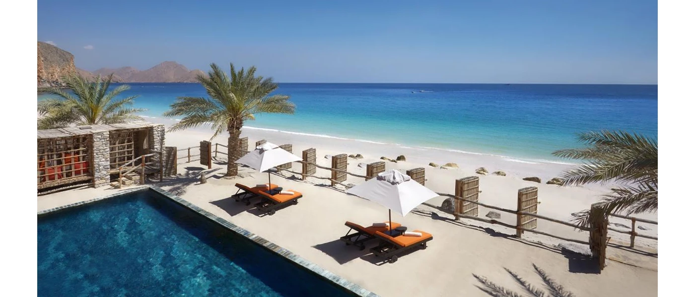 A white-sand beach with orange loungers, in front of a swimming pool and palm trees