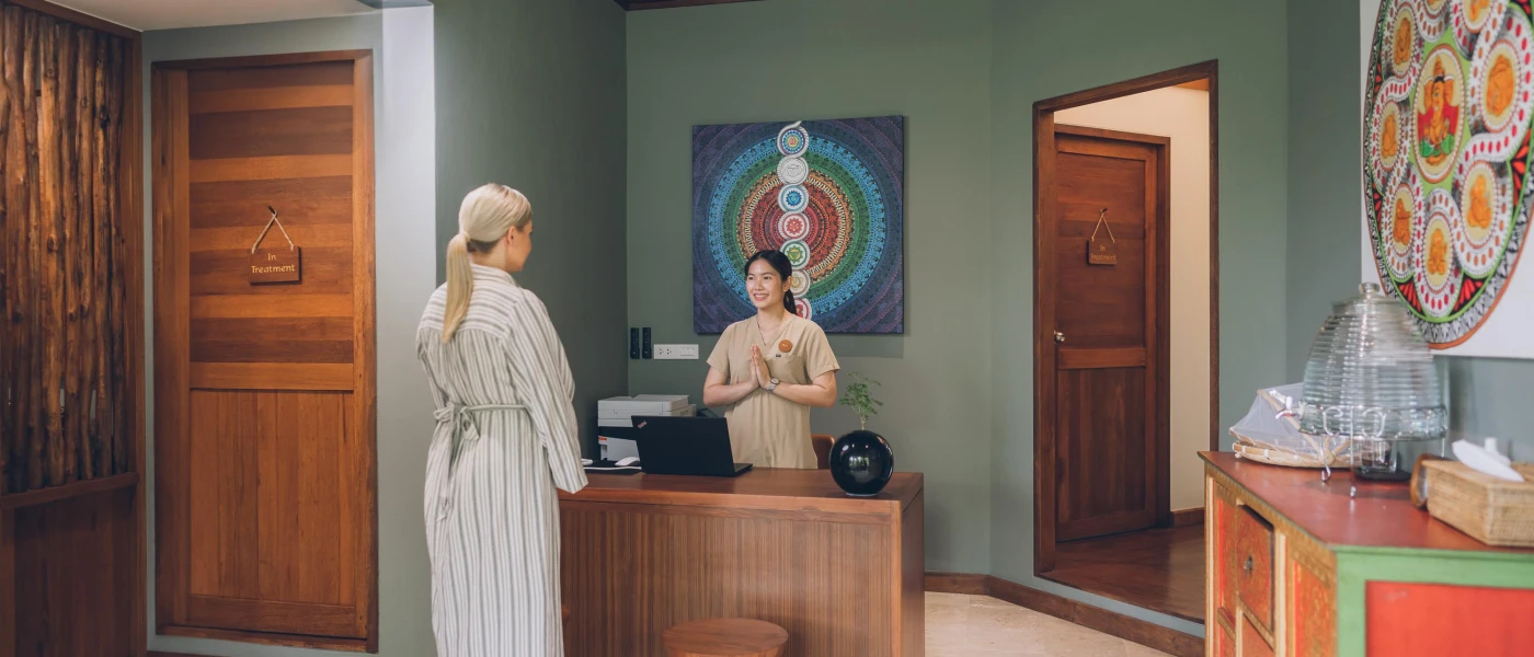 Woman in a striped robe in greeted by a smiling staff member in front of a picture depicting the chakras in a room with wooden doors