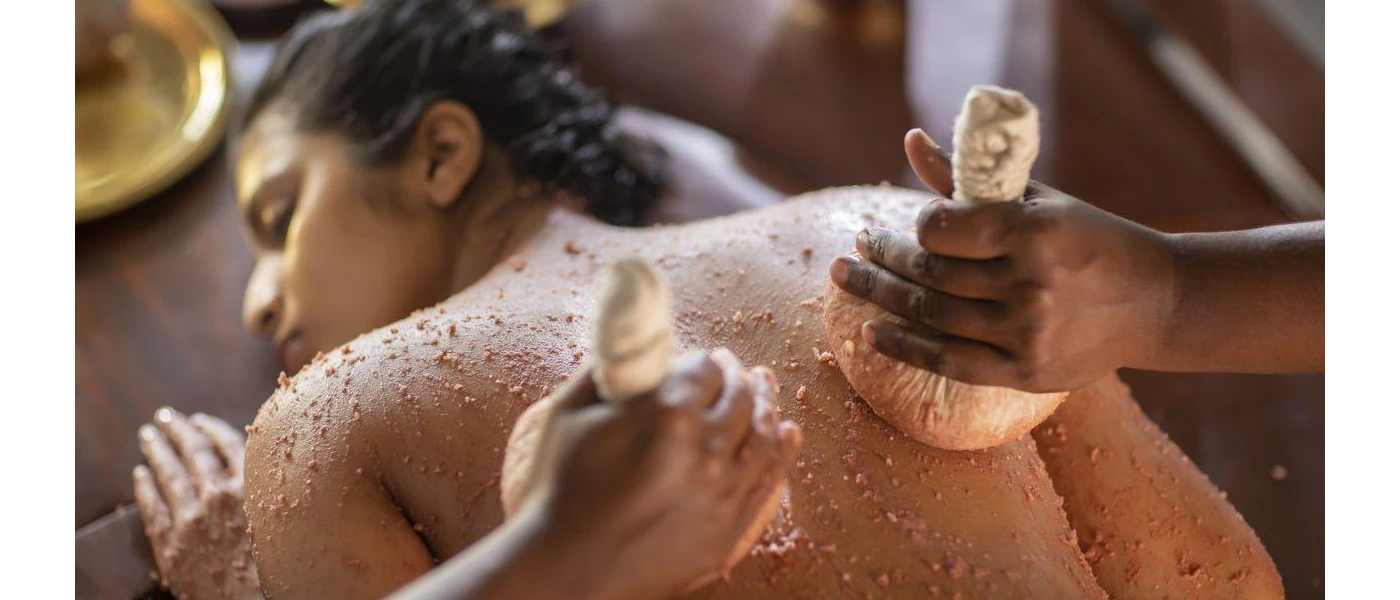Woman lies on her back with her eyes closed as two therapists exfoliate her skin