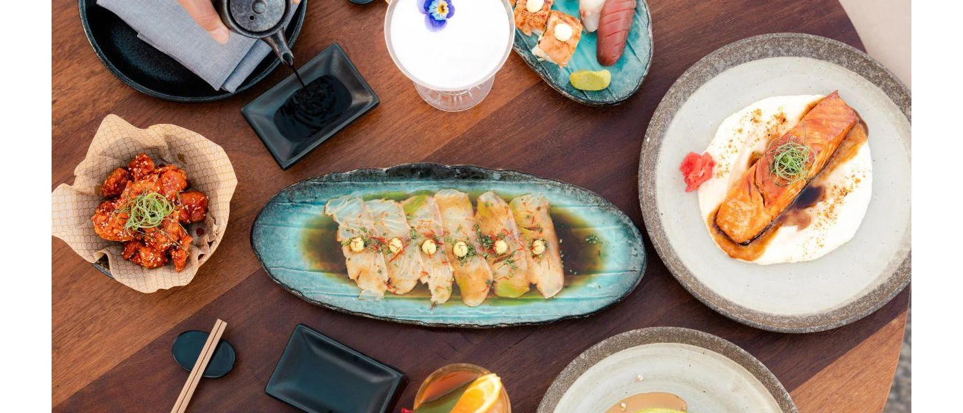 Table laid with plates of seafood including a fillet salmon, sliced tuna and sushi, and a cocktail glass topped with white liquid and a purple edible flower