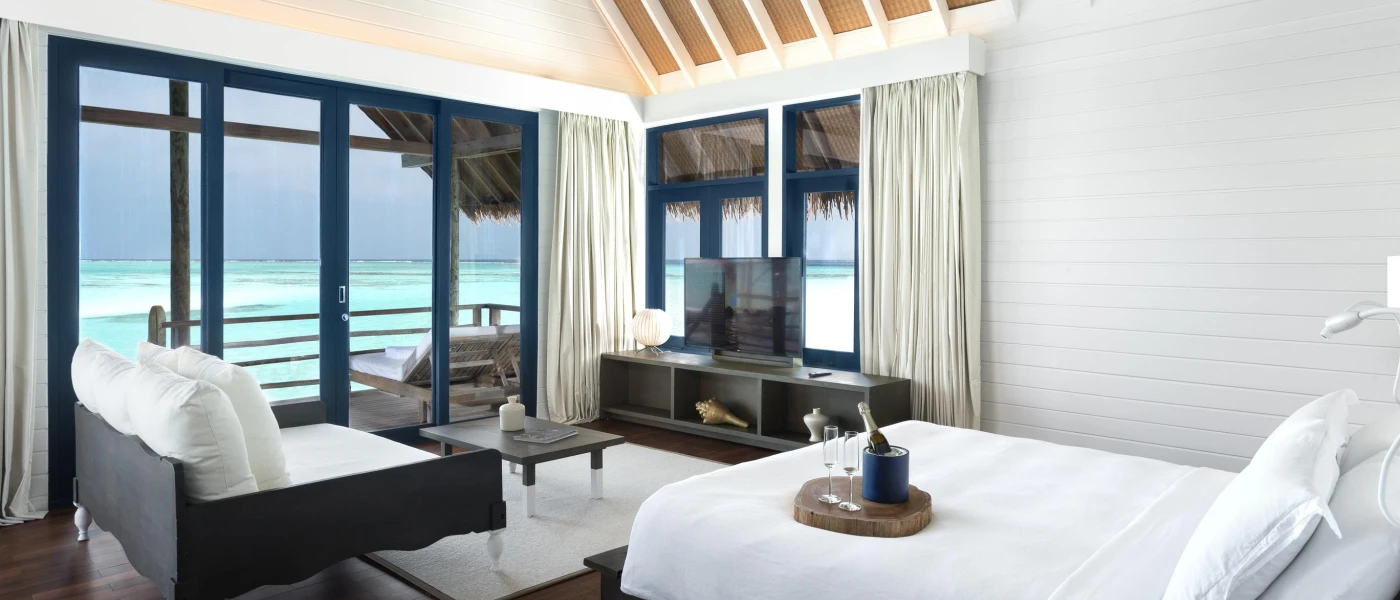 Open-plan villa bedroom and living area with white bedding and sofa, dark-wood furniture and full-length doors with a view of the ocean
