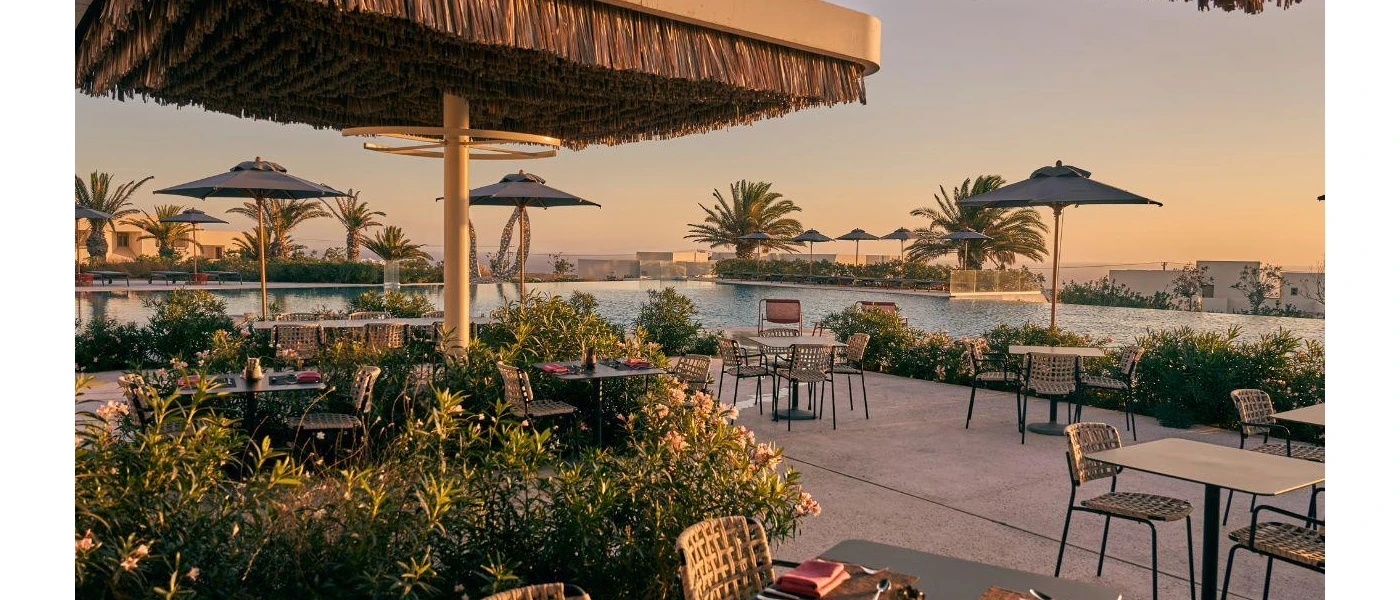 Lake-like swimming pool at sunset, surrounded by palms and shrubs, grey parasols and a terrace with tables, chairs and a thatched parasol
