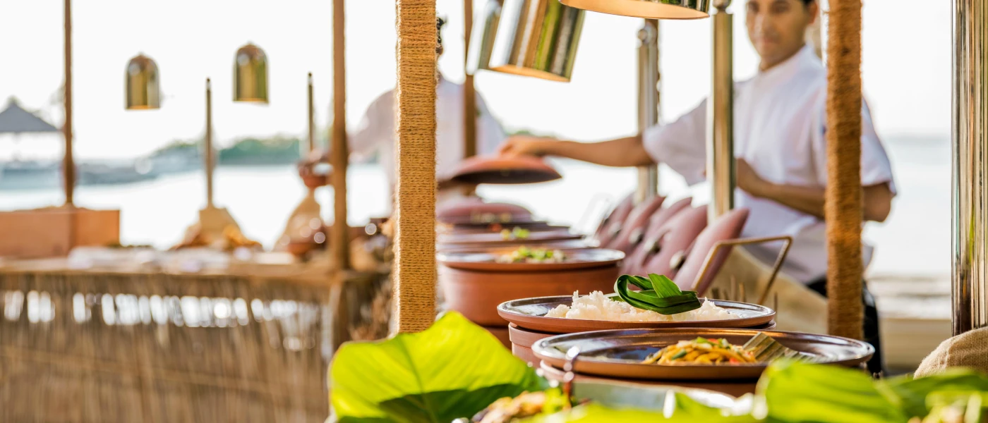 Pots of food lined up buffet-style with a chef in whites behind, and the ocean in the background