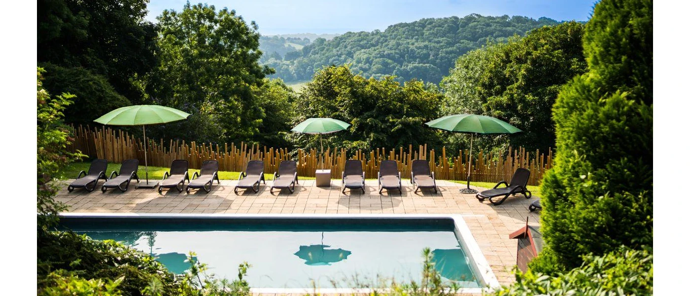 Outdoor swimming pool in a sunny terrace, surrounded by loungers, green umbrellas and greenery