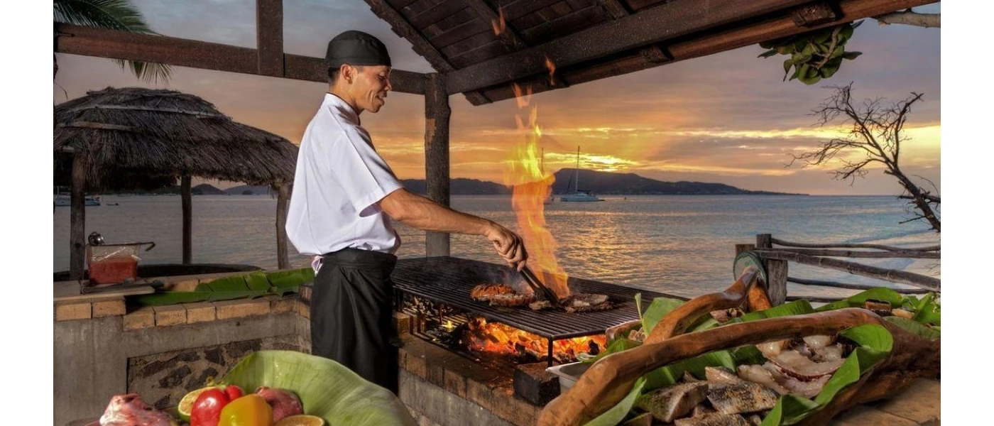 Chef in an open-air kitchen cooking over an open flame surrounded by ingredients and overlooking the sea