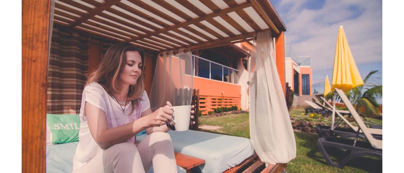 Smiling woman in white looks down at her mug while sitting on a wooden daybed