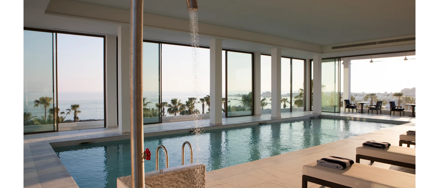 Indoor pool with floor-to-ceiling windows, white-cushioned loungers with striped towels, all overlooking the sea