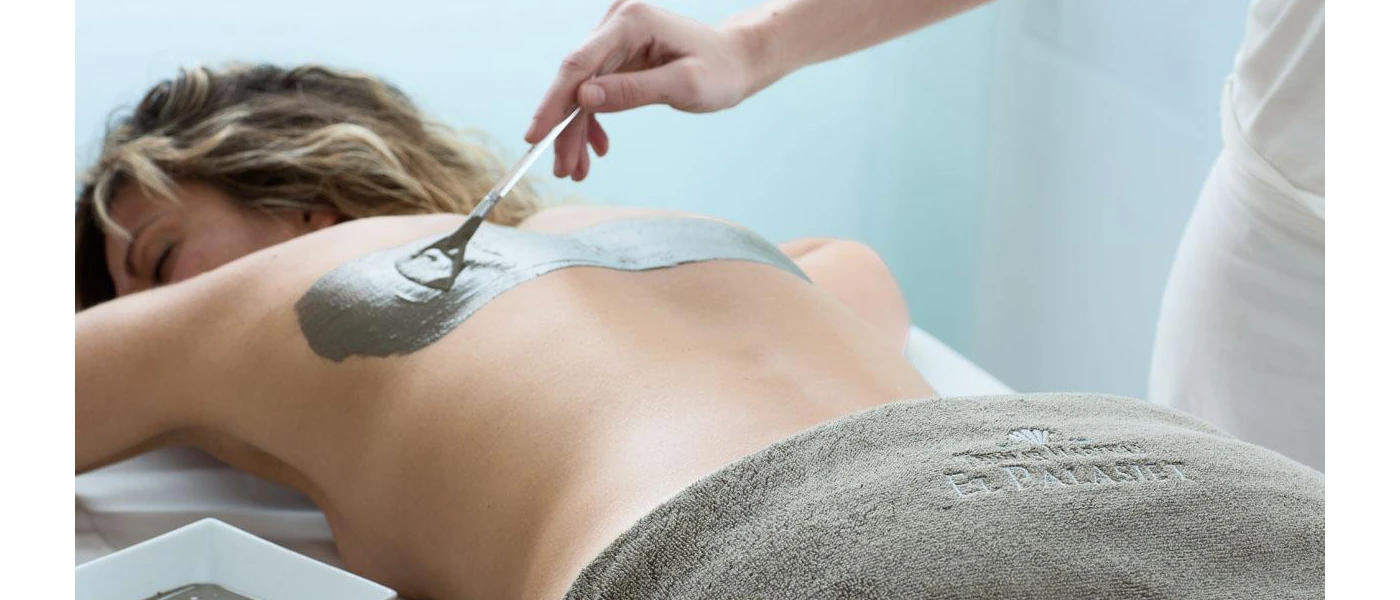 Woman lies on her front with a grey towel partially covering her, as a therapist spreads a mud-like substance on her back