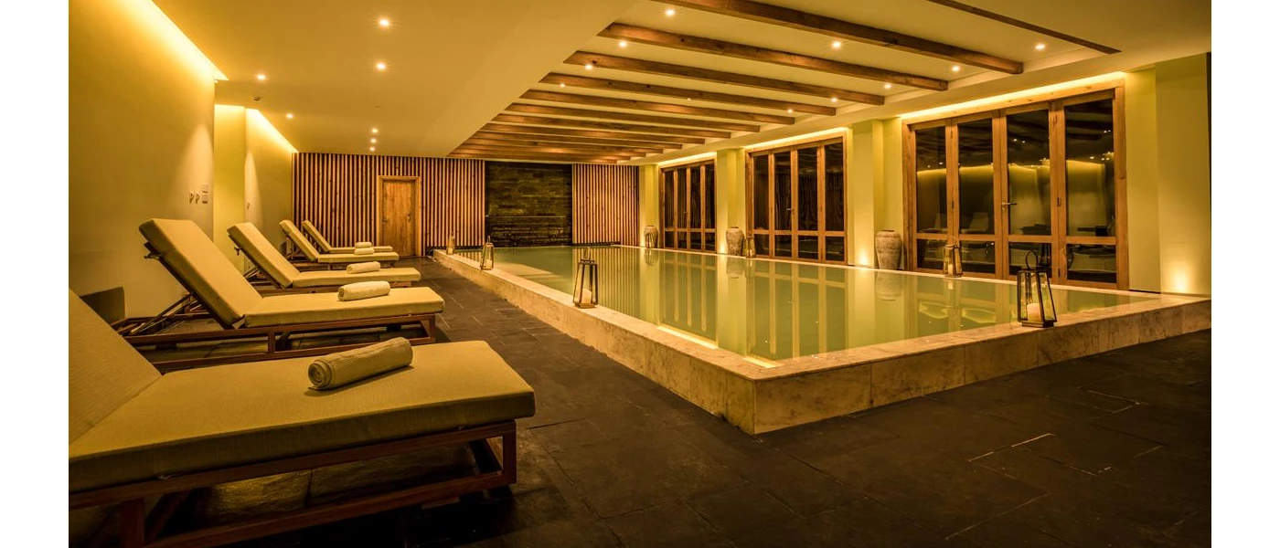 Lantern-lit indoor pool surrounded by cushioned wooden loungers and with wooden beams in the ceiling