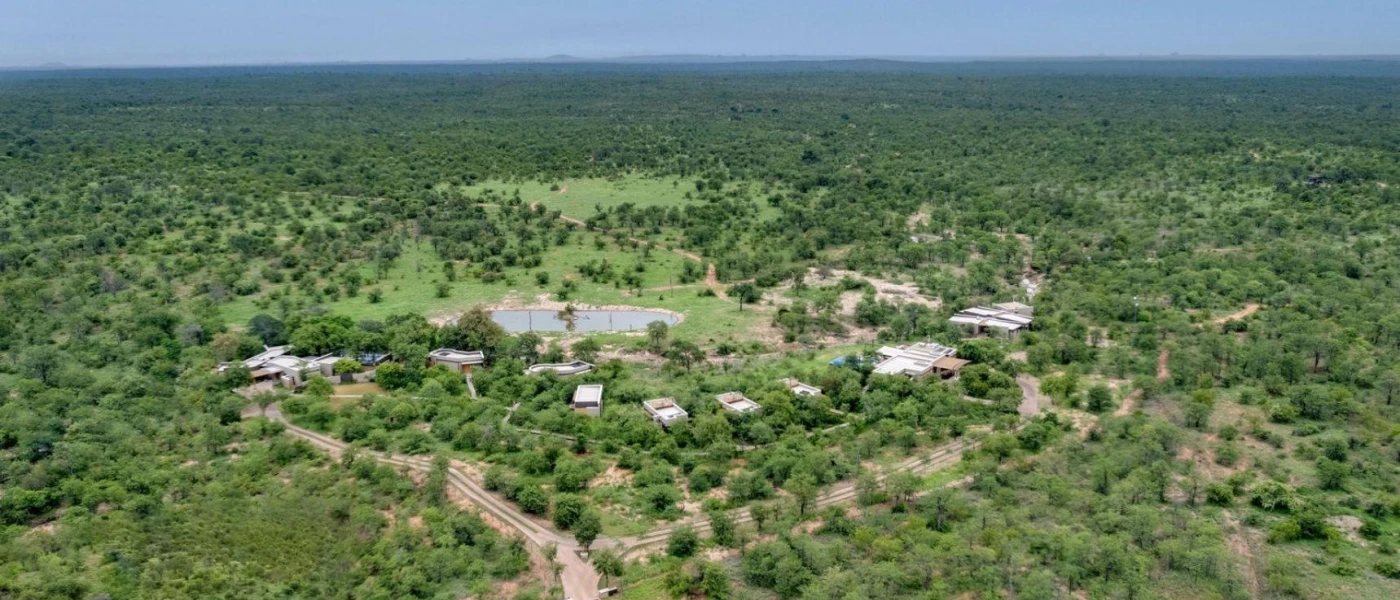 Green wilderness under a blue sky, with sand-coloured lodges peering out from the greenery and a waterhole in the background