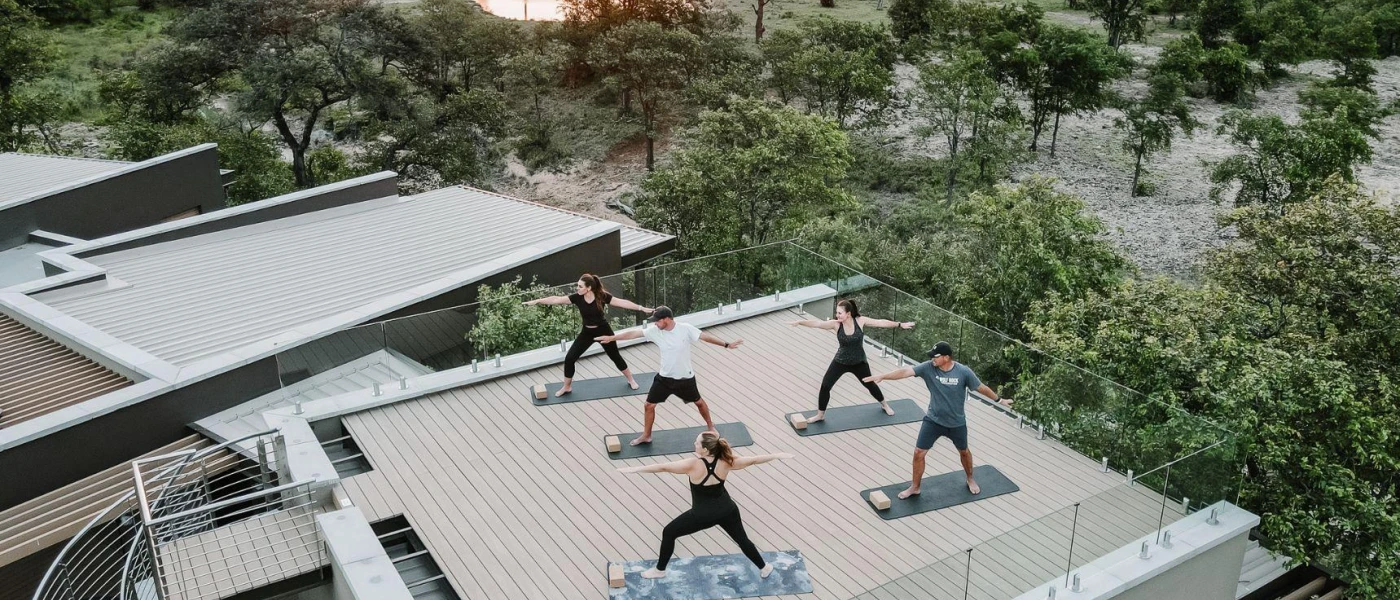 Group in active wear take part in a yoga class on a wood-decked roof terrace surrounded by reserve greenery