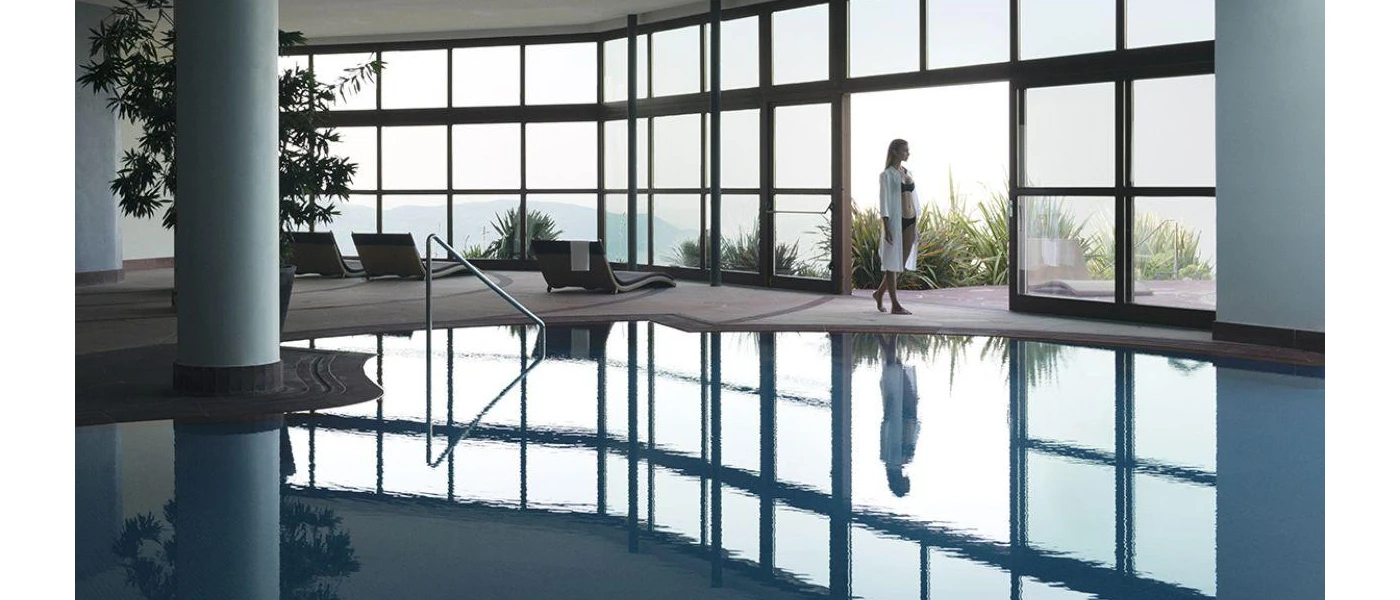 Woman in a black bikini standing next to an indoor pool with floor-to-ceiling windows