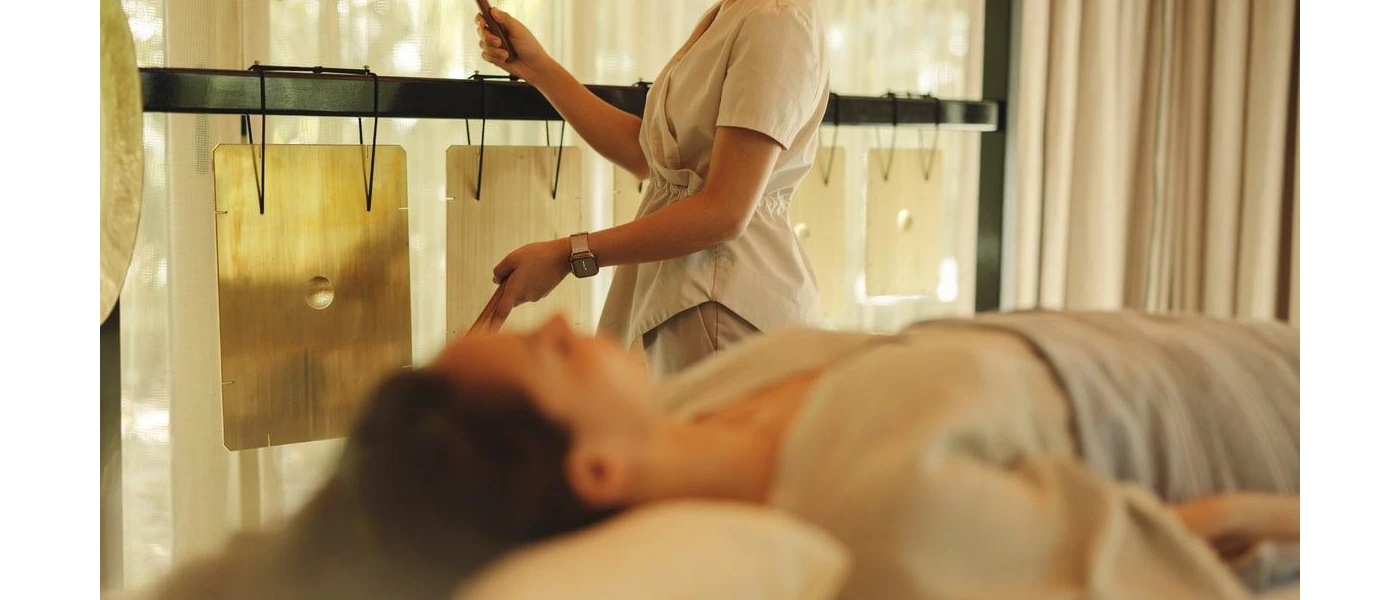 Woman with long dark hair relaxing on a massage bed as a therapist practices sound healing next to her