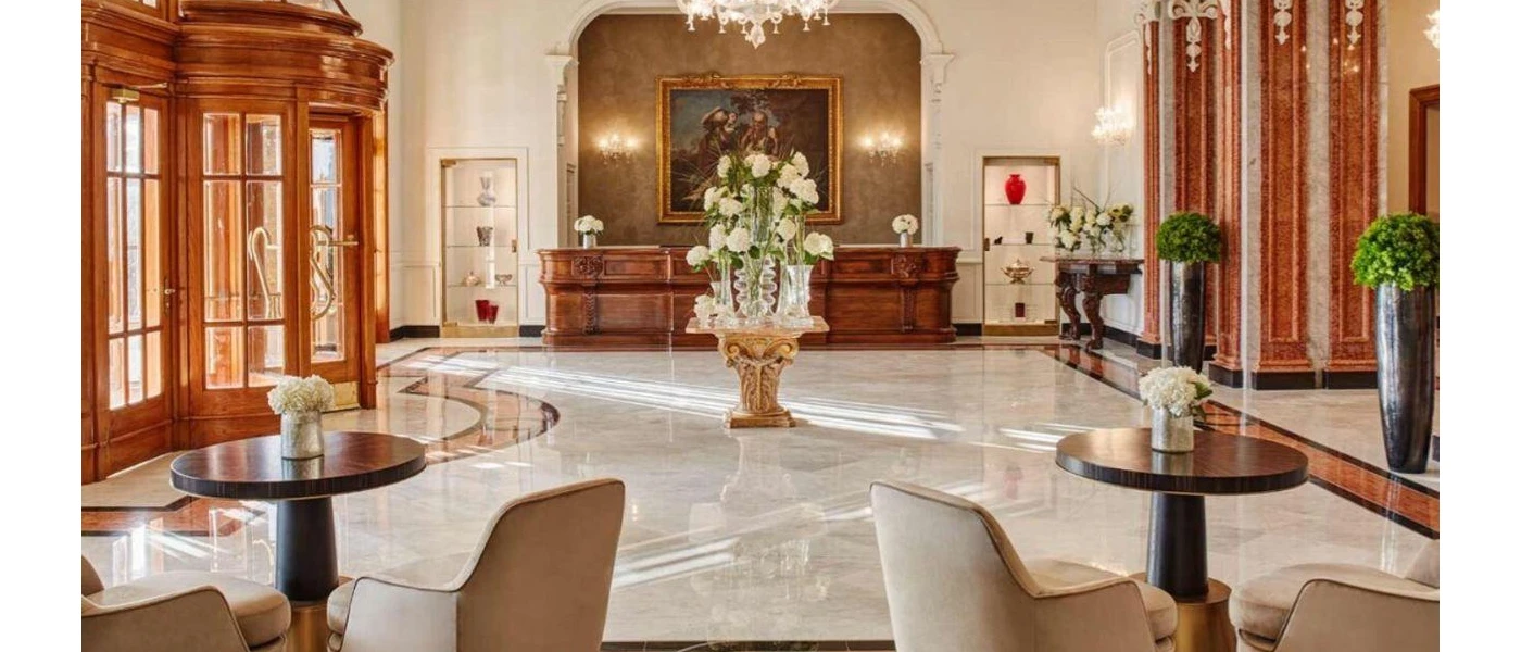 Reception area with marble floors, wooden revolving doors, a central wooden table with a large vase of white flowers, a wooden desk and a painting with an ornate gold frame hung behind it