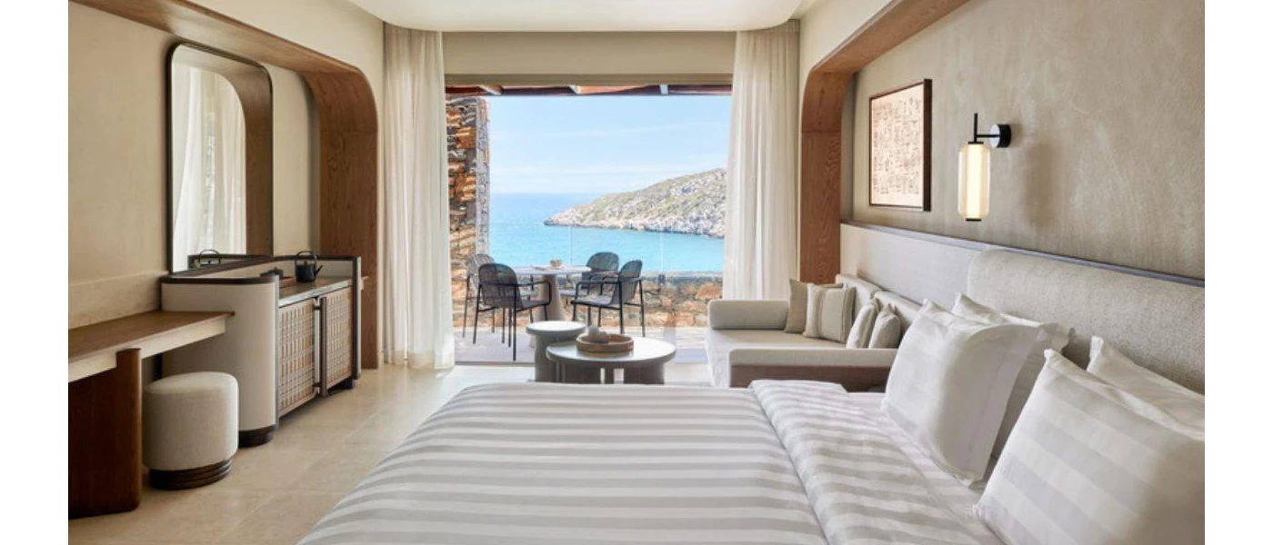 Neutral hotel room with striped bedding, lantern-like wall lights, a wooden dressing table and stall and a furnished balcony with sea and hillside views