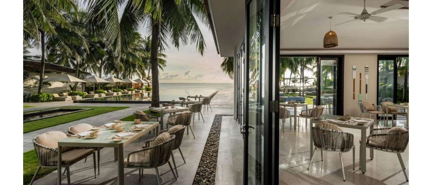 Modern restaurant with indoor and outdoor tables, full-length glass and a view of the ocean and palm trees