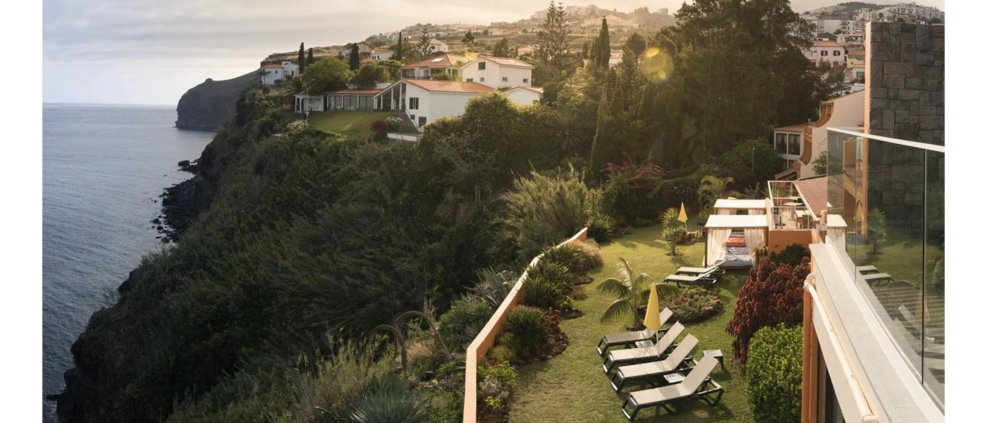 Lawned terrace with four-poster daybeds with white curtains and wooden loungers, overlooking a belt of greenery and the sea