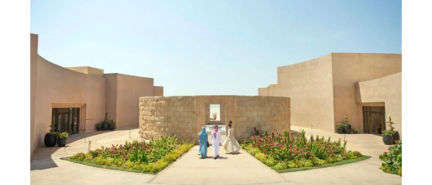 Three women walk through a sunny courtyard surrounded by sand-coloured cube-like buildings, with flower beds leading to a central water feature flanked by stone walls
