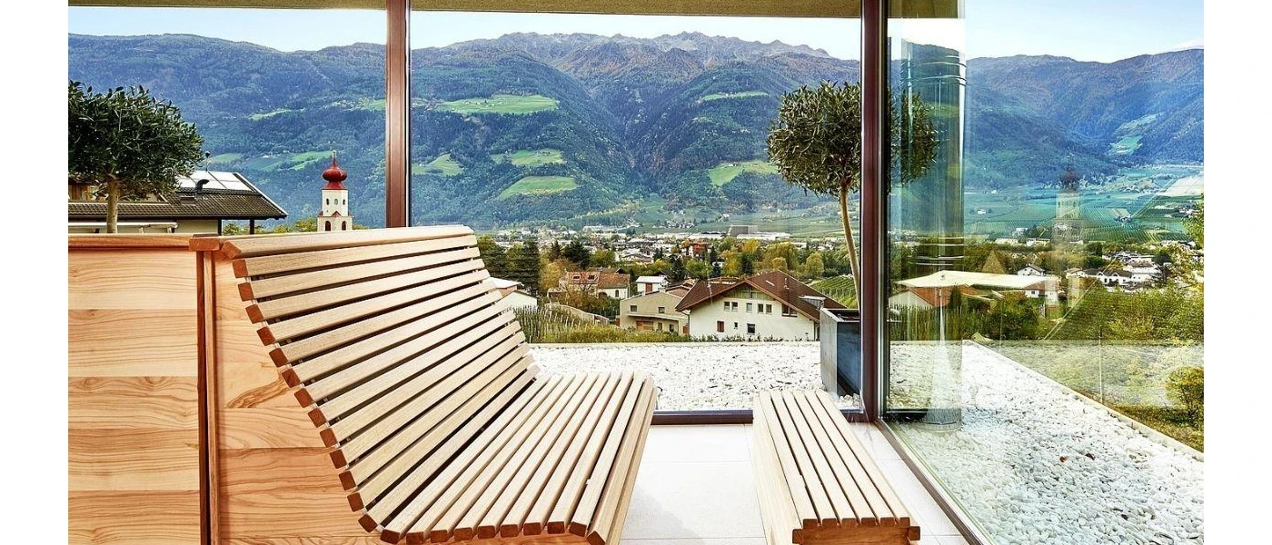 Wooden bench in a room with panoramic glass windows and views of a village below, and surrounding mountain peaks