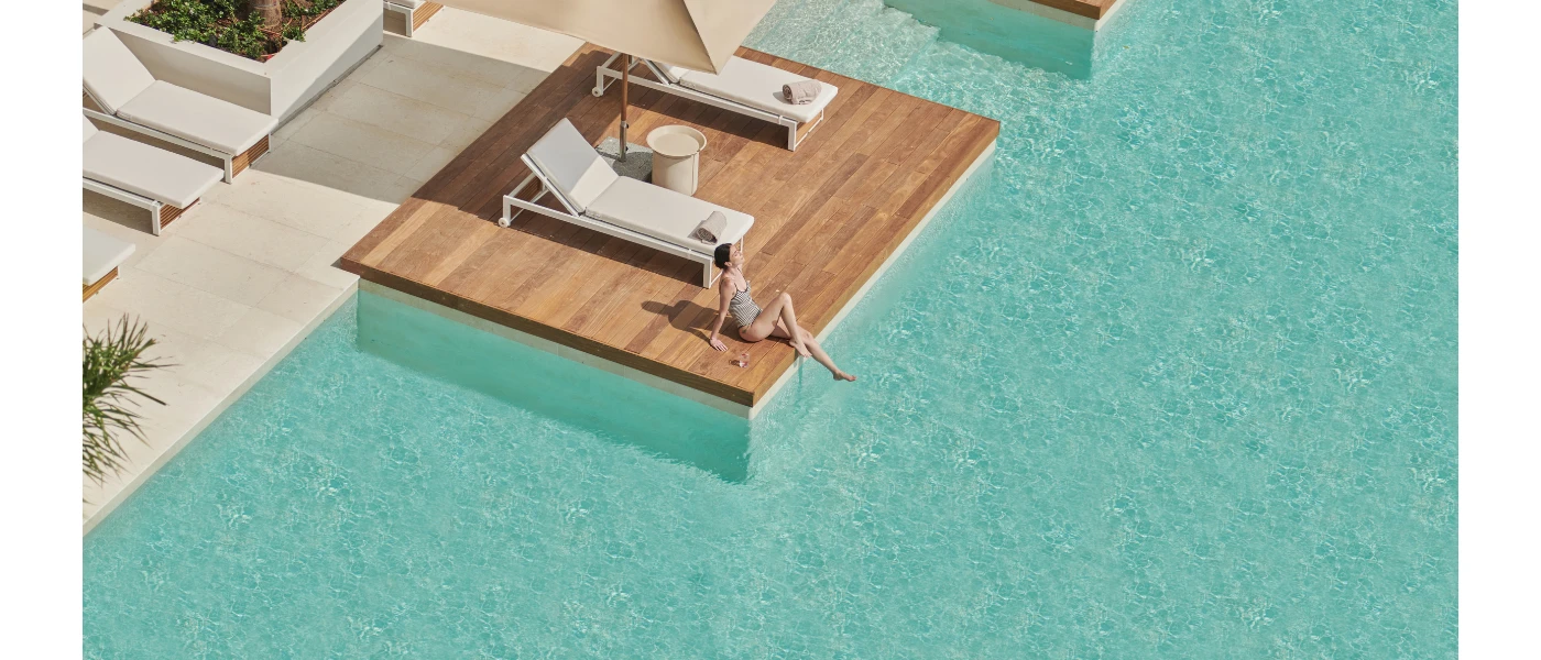 Woman in a grey swimsuit dangles one leg in a swimming pool while sitting on a wooden terrace furnished with ivory-cushioned loungers