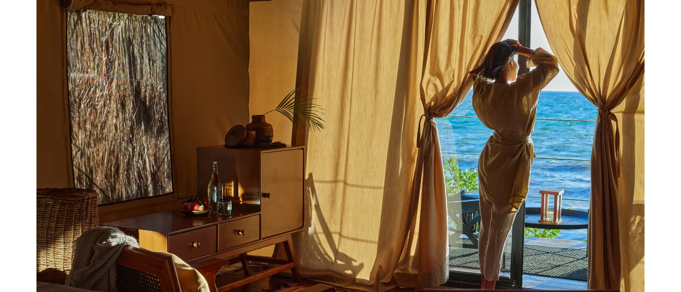 Woman stands in a robe looking out to sea from her terrace windows, with ivory drapes cascading from the walls and wooden furniture inside