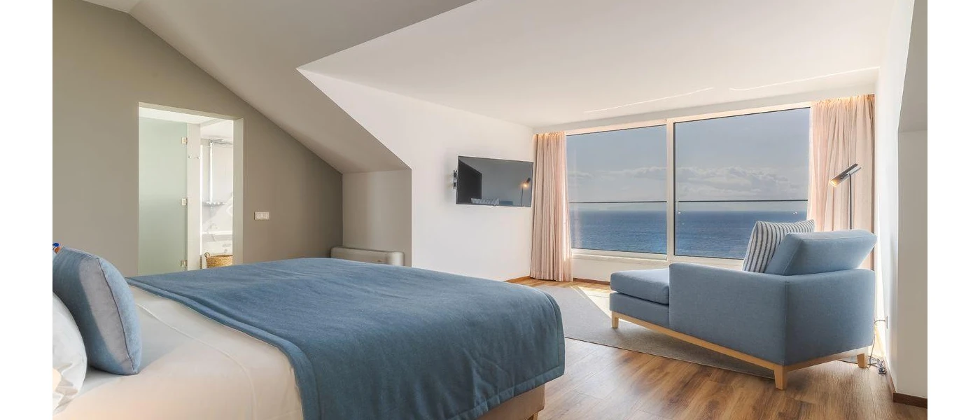 Modern hotel room with white bedding and a blue throw and cushions, a blue chaise longue and a large window overlooking the sea
