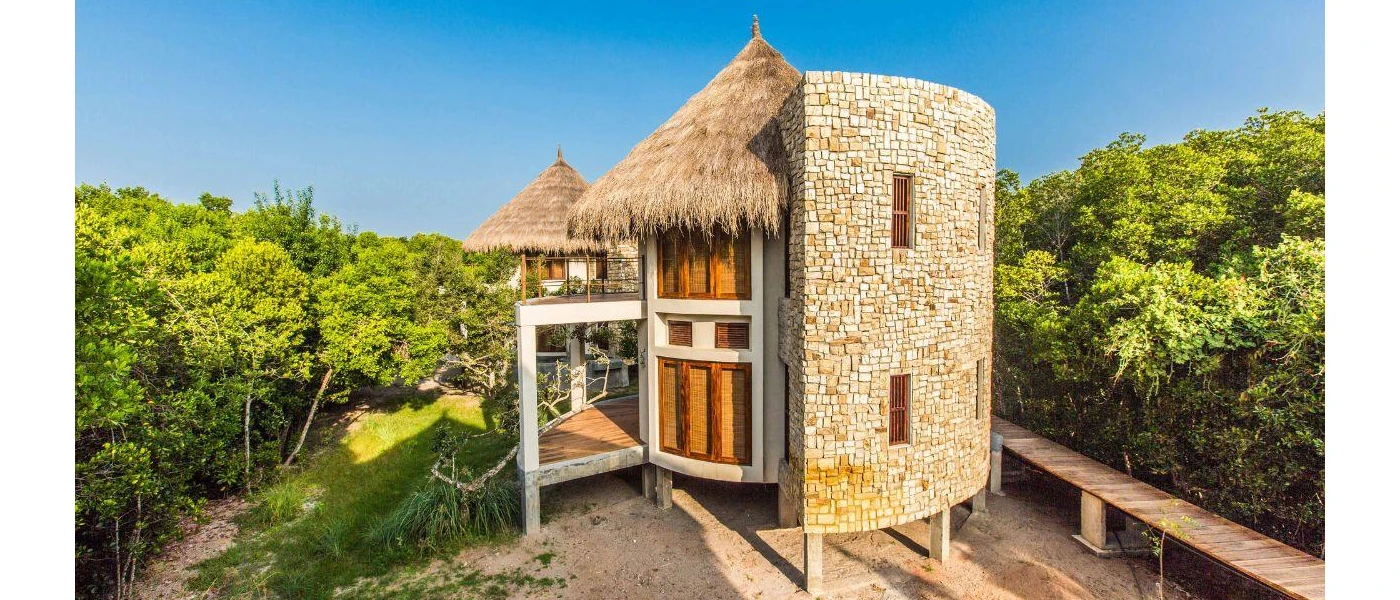 Circular stone-walled building with a triangular thatched roof, surrounded by a forest