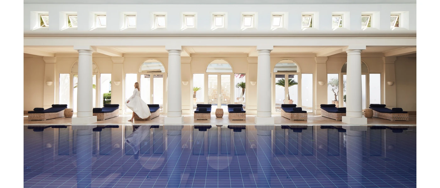 White-robed woman walks past an indoor pool surrounded by white pillars and rattan loungers with navy cushions