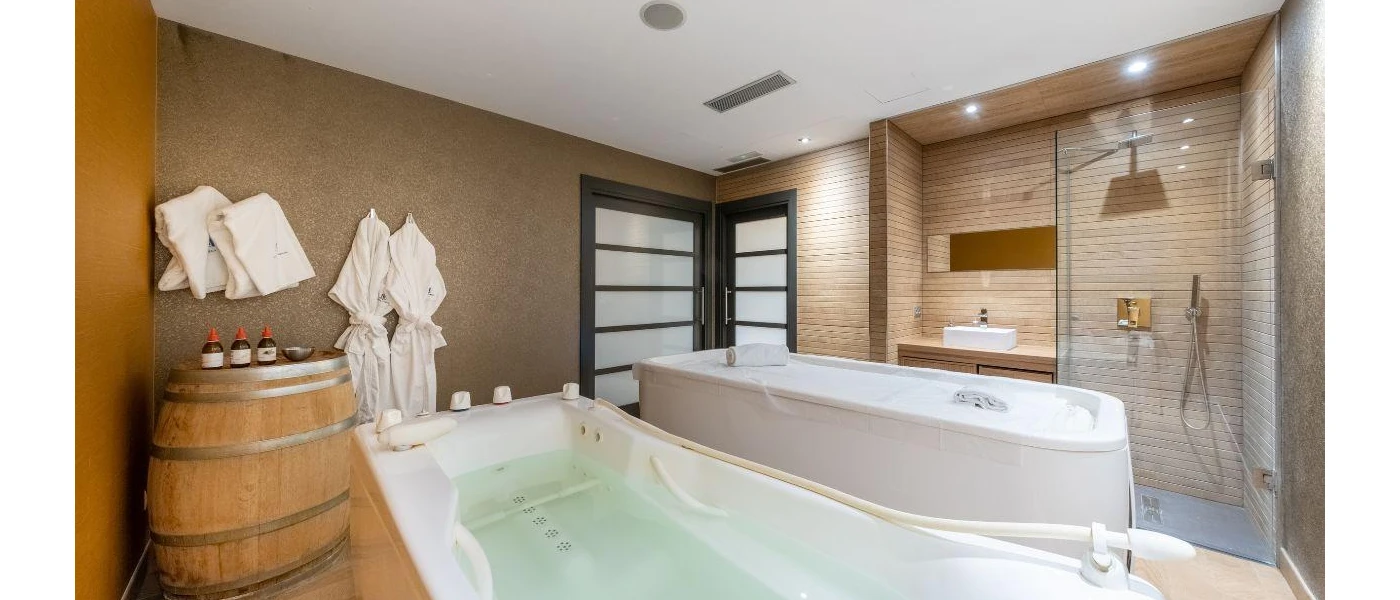 Spa treatment room with a shower, bath and massage bed, a wooden barrel with bottles and white towels and robes hung on the wall