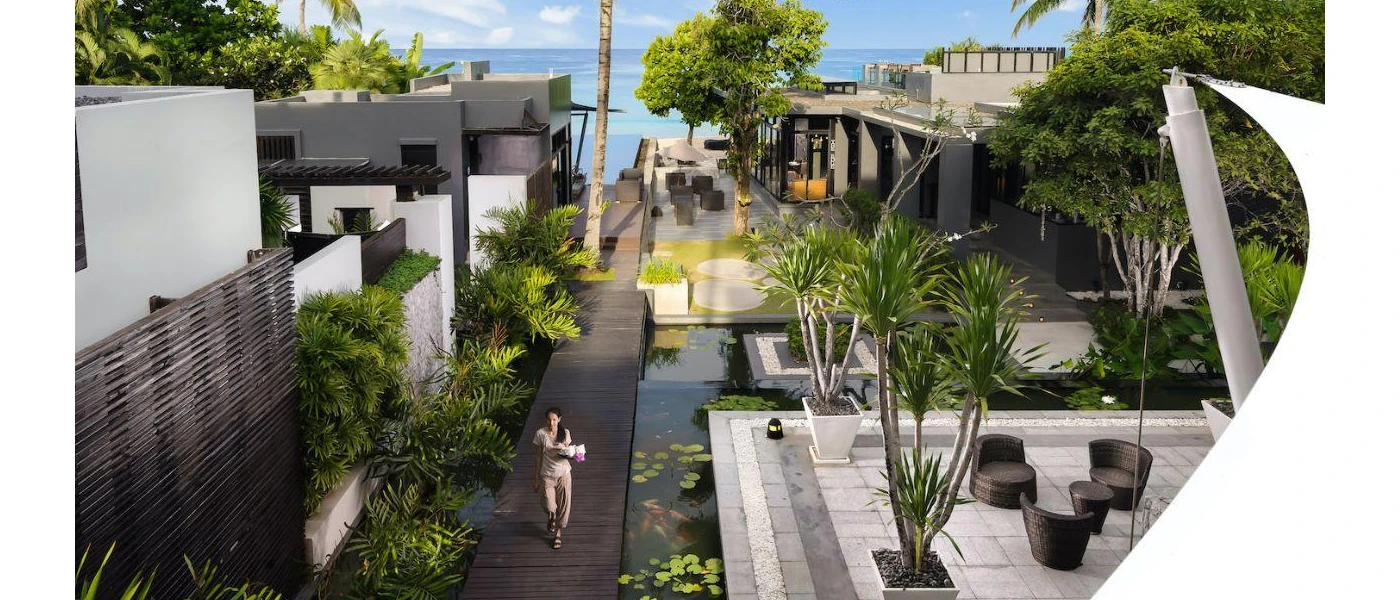 Woman in cream walks down a dark-wood walkway surrounded by villas, palm trees and ponds of lily pads