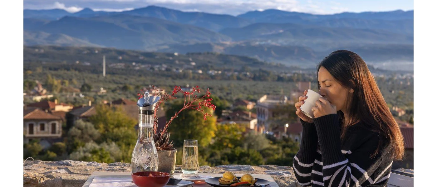 Woman in a striped jumper sips from a white cup with a plate of eggs and hollandaise sauce in front of her, with mountains and a resort beneath her