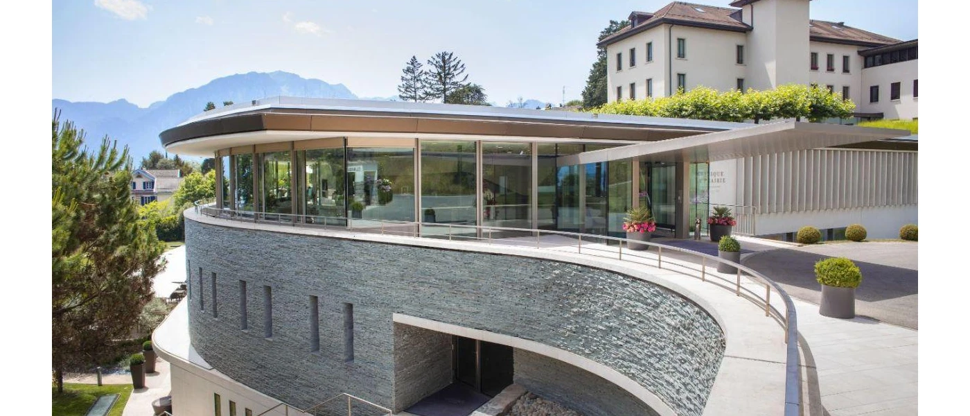 Curved balcony area next to a room with floor-to-ceiling windows, with mountains and multi-storey white buildings in the background