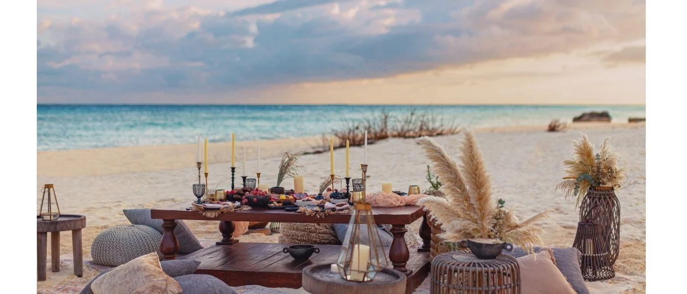 Dark-wood low coffee table on the beach set up for a picnic, with black and brass candlesticks, bowls of fruit, bean bags and surrounding wooden stalls with lanterns