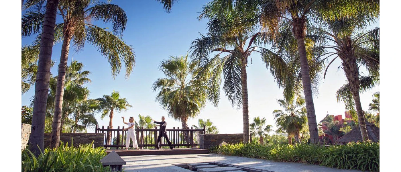 Two people practice Tai Chi in a tropical garden under a blue sky