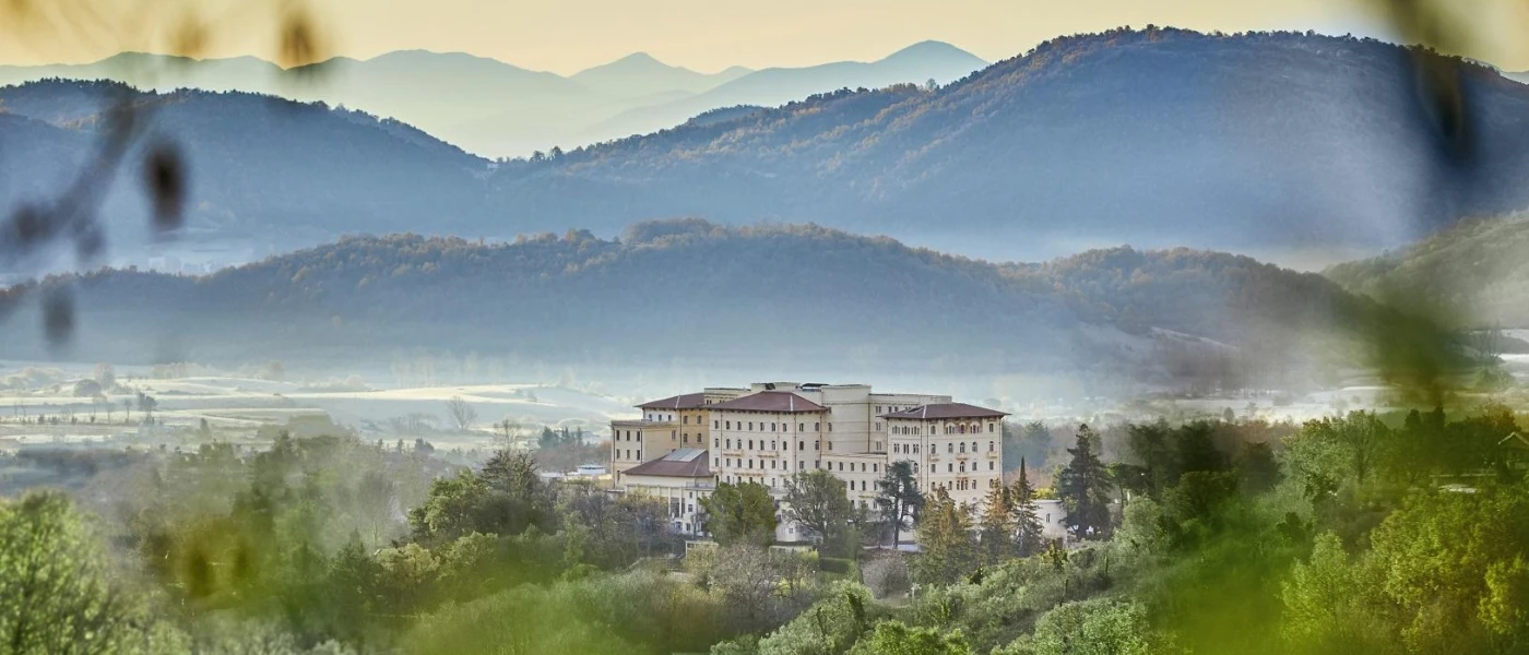 Palazzo Fiuggi exterior, with multiple storeys and sand coloured buildings, tucked among the countryside surrounded by misty peaks