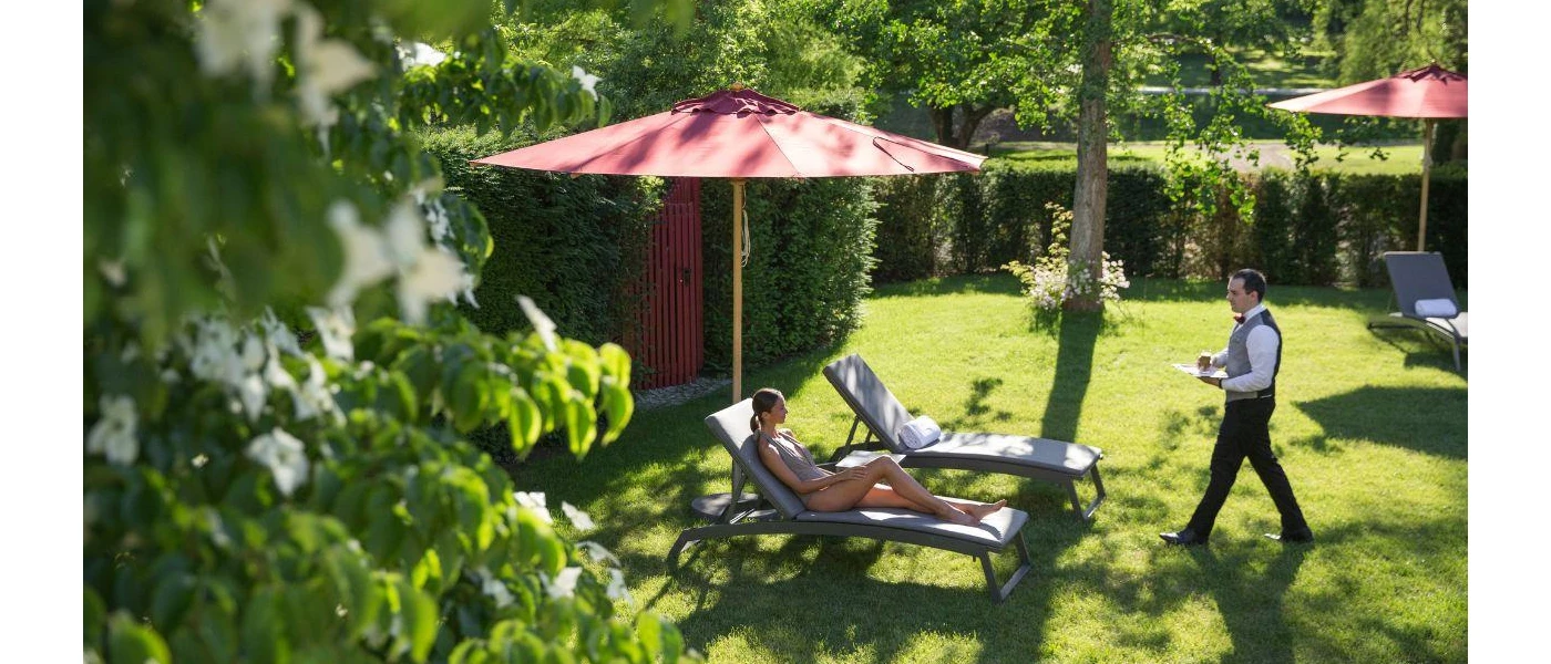 Man in waistcoat and bow tie carries a tray over to a woman lying on a sunbed in a swimsuit in a sunny garden
