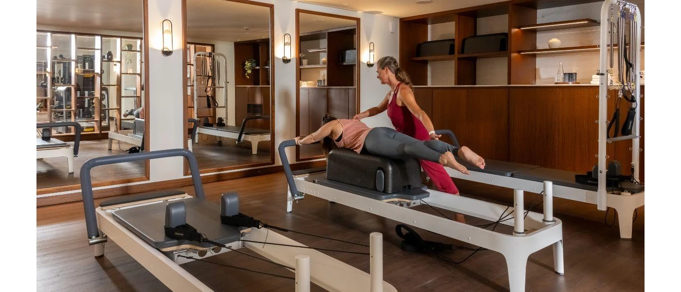 Woman lies on her front on a reformer Pilates machine as a staff member in red active wear supports her posture