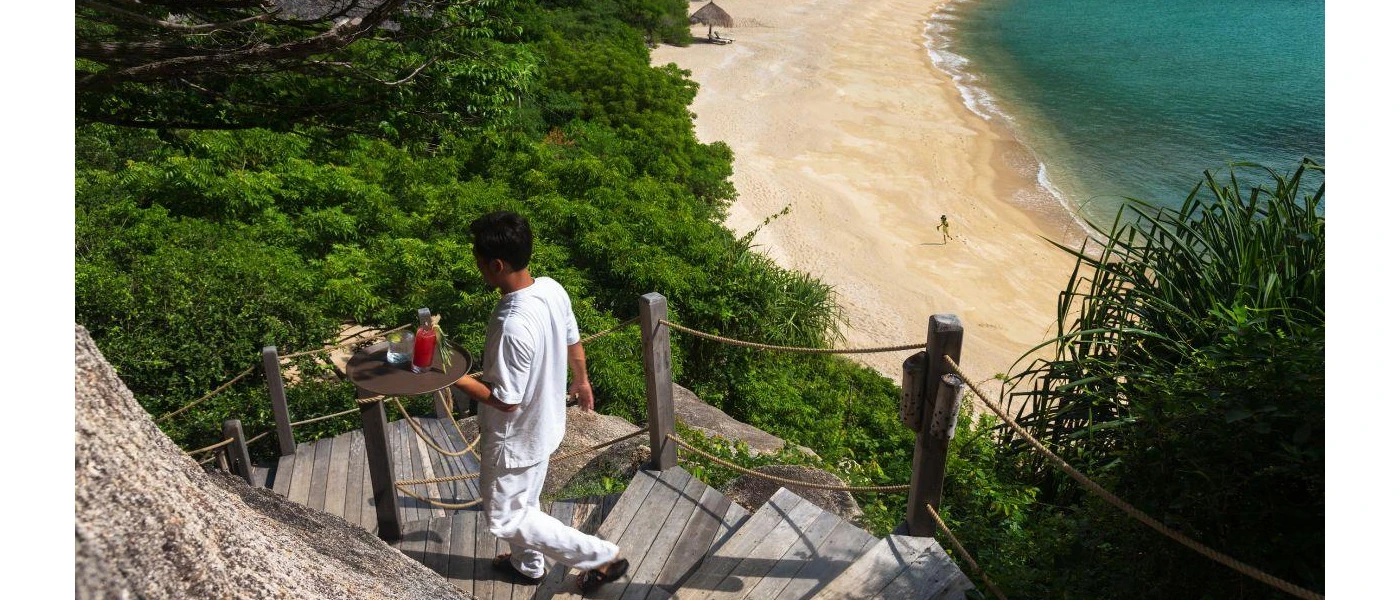 Staff member walks down a curved hillside staircase carrying a tray, as the waves lap a golden sandy beach below