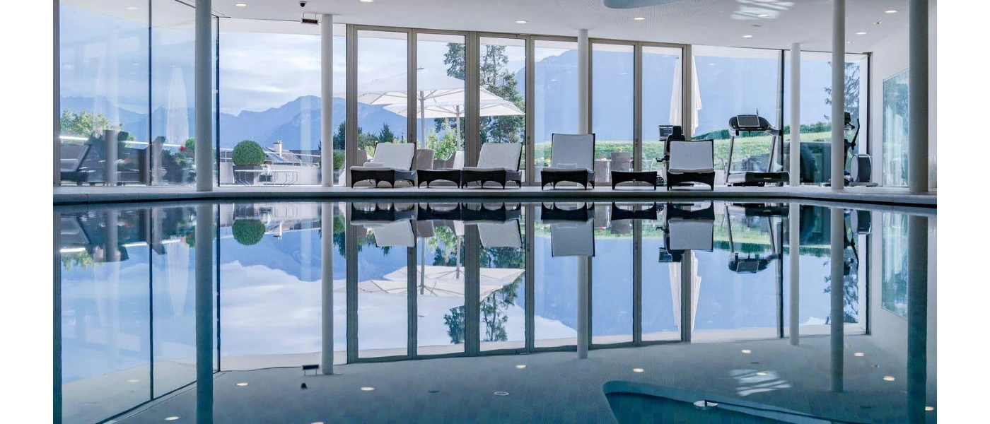 Indoor pool surrounded by white-cushioned loungers with floor-to-ceiling windows showcasing the mountains beyond
