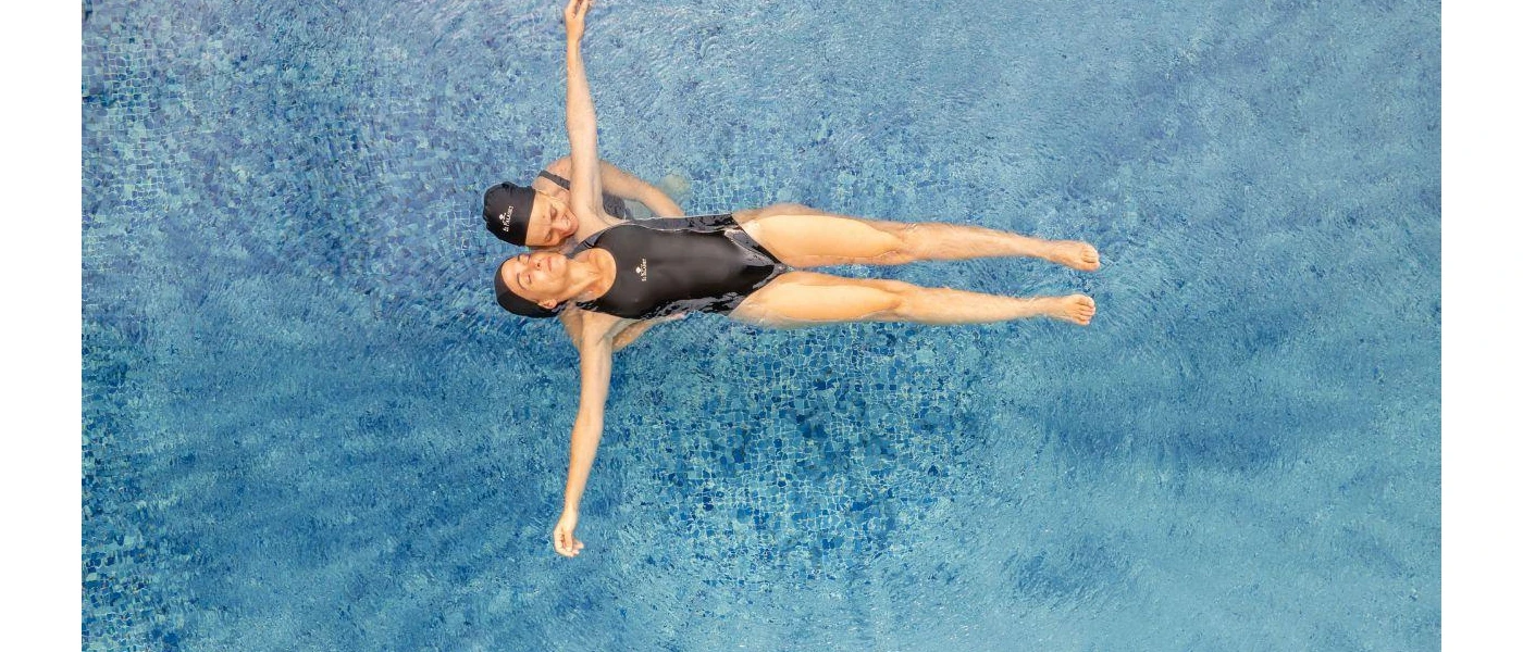 Woman in a black swimsuit and swim hat floats on her back in a pool, supported by another woman
