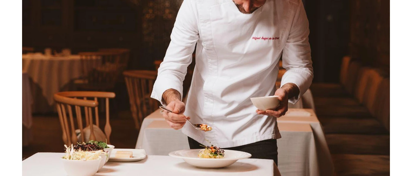 Man in chef whites spooning food onto a white plate, on a table with a white tablecloth