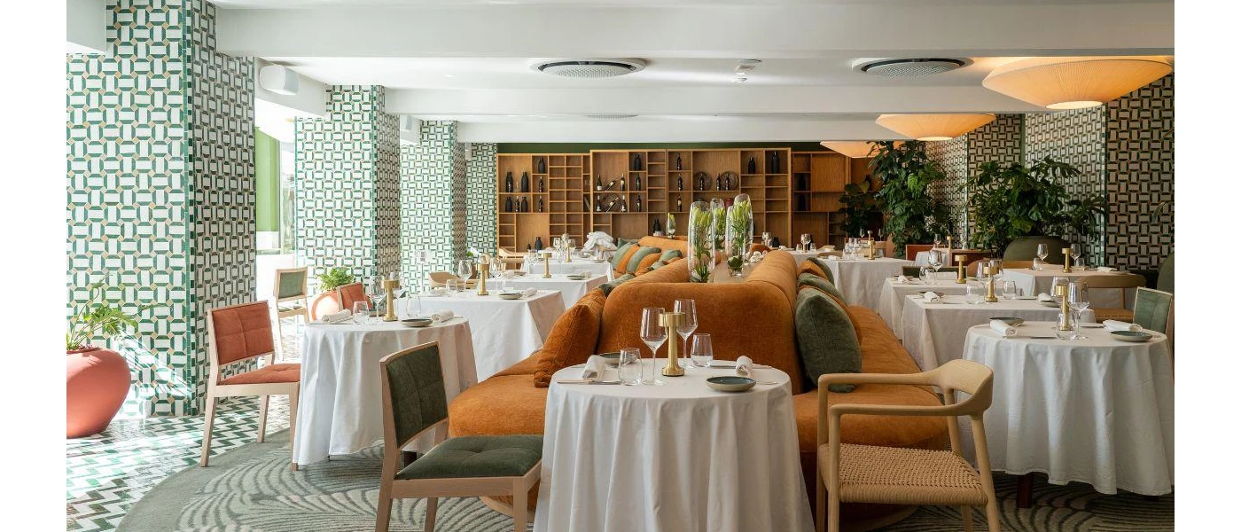 Restaurant with white tablecloths, an orange sofa centrepiece for seating, and a shelving unit full of wine bottles
