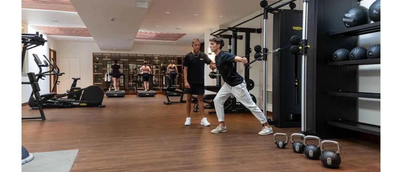 Man in a black t shirt and grey tracksuit bottoms uses a weights machine in a gym while a PT looks on