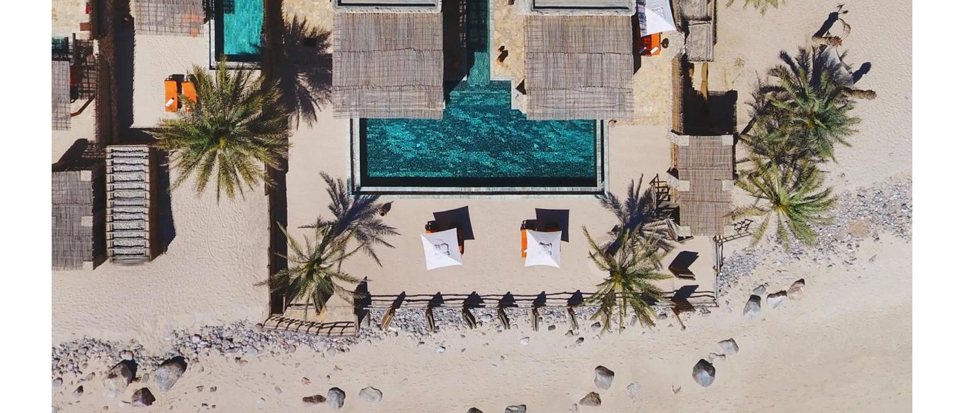 Aerial view of Six Senses Zighy Bay, including grey rooftops, palm trees, an infinity pool and orange loungers, all set in ivory sands