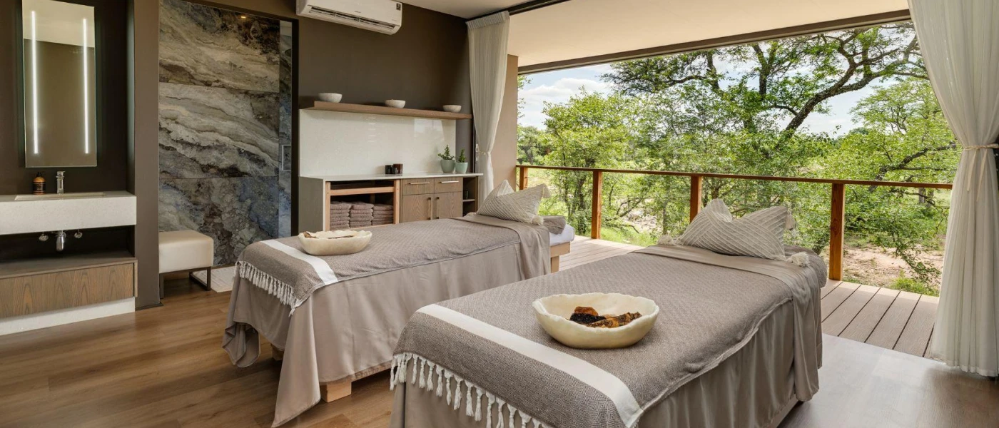 Luxe spa treatment room with two beds with grey fabric coverings, a marble-look wall, vanity unit and mirror with LED lighting and doors open onto a balcony with reserve views