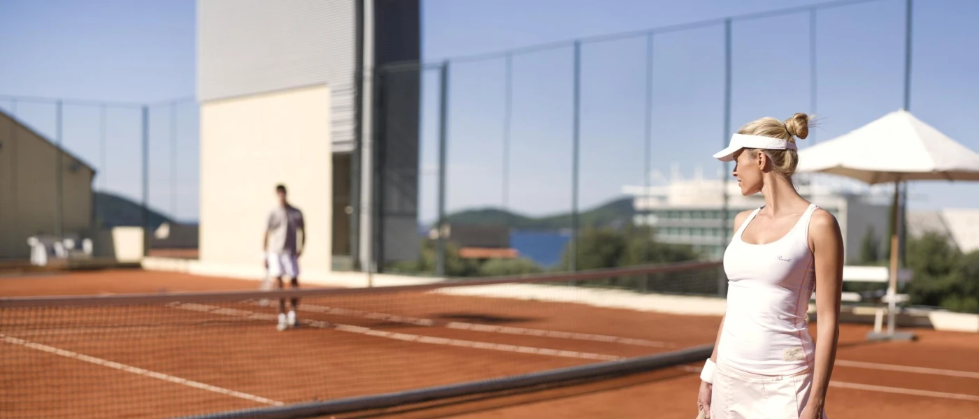 Woman in tennis whites and a visor standing on a court looking over the net to a male playing partner, slightly blurred in the background