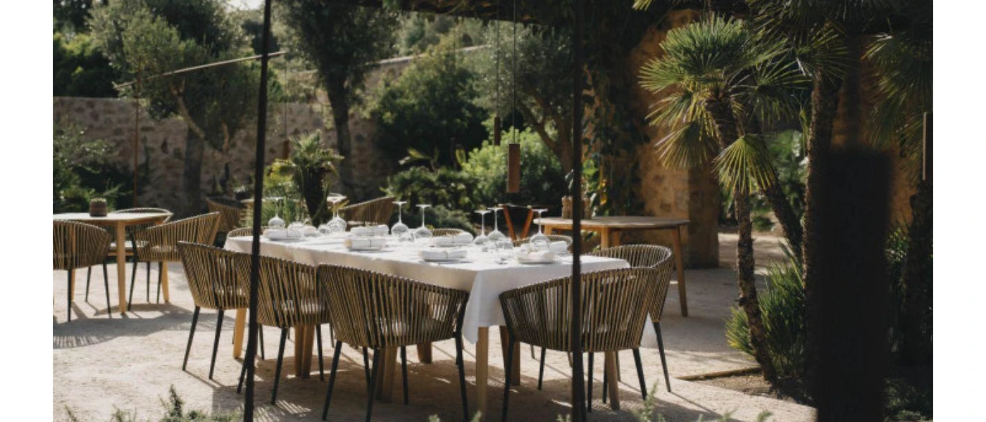Table with white table cloth and wooden seats laid out for a meal in a lush green courtyard